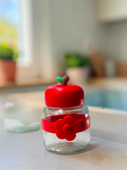 Miso station apple-shaped glass storage jars with colorful lids and flower-accented silicone bands on a white surface.