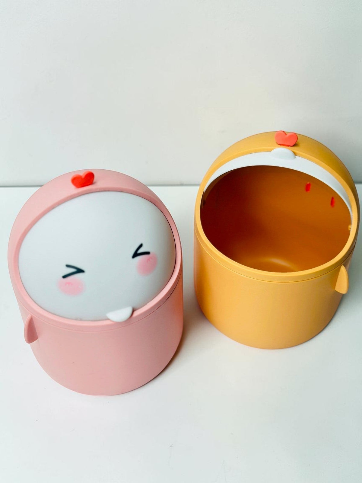 Miso station cute character desktop waste bins in pink and yellow with swing-top lids on a white brick background.