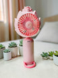 Miso station blush pink lotus-inspired portable handheld fan with a bottom loop, sitting on a desk with succulents.