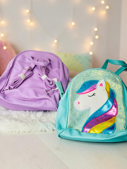 Miso station unicorn mini backpacks in purple and glittery teal with multi-colored manes on a white brick background.